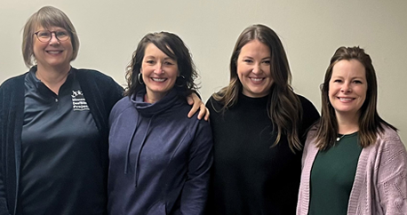 All four are white women standing closely together in front of a white wall and smiling at the camera. They are all wearing dark shirts and with one pink sweater.