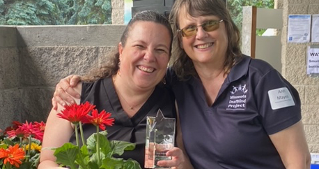 Two white women stand close together. On the left is Amy who is holding a Shining Star glass award and flowers. Next to her is Ann Mayes.