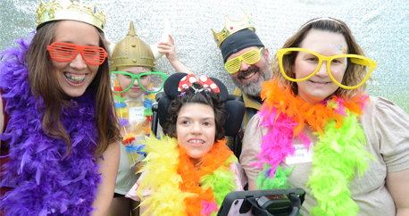 3 light skinned adults and two children are wearing silly glasses, hats and colorful boas in a photo booth. One child sits in her wheelchair and has a high tech communication device in front of her.