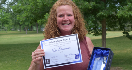 A white woman with curly red hair holds up a Shining Star certificate and glass award.