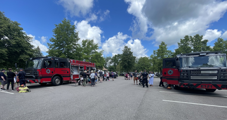 Two fire trucks are parked in a lot with many people milling between them speaking with fire fighters and experiencing different parts of the fire equipment.