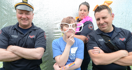 A white teenage girl wears oversized, silly white glasses and has her hand on her chin. Around her are three firefighters with their arms crossed, wearing silly costume hats and smiling at the camera.