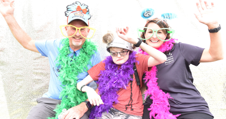 Dan, Danny and Deanna Rothbauer are wearing colorful boa, silly hats and have their arms up in celebration in a photo booth.