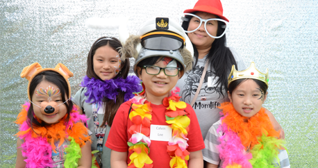 An Asian American Mom and her four children are wearing silly glasses, hats, and colorful boas in a photo booth.