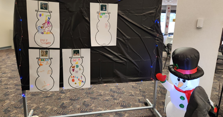 Image taken in a conference room. A standing board is covered in black paper and has twinkle lights around it. There are pictures of snowmen decorated by deafblind children/youth. An inflatable snowman is to the right.