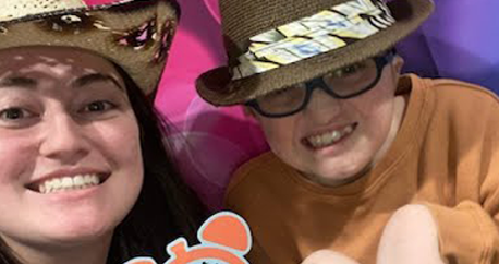 a pre-teen white boy and a young white woman with dark hair are both wearing silly hats, smiling, and standing in front of a colorful backdrop.