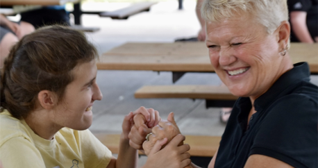 A teenage girl with brown hair in a braid has her hands resting over the hands of a middle-aged woman with short blond hair who is using tactile ASL.