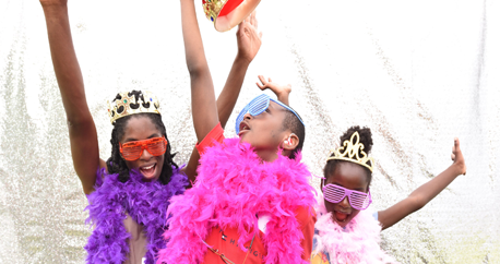 An African mom and her son and daughter are dressed up in colorful boas, glasses and crowns and throwing their hands in the air in fun.