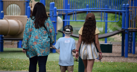 A mother with long dark hair, her young son with very light hair and teenage daughter with long brown hair hold hands and walk towards a playground together.