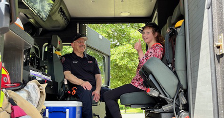 A white teenage girl is sitting inside of the fire truck giving a thumbs up. Next to her is a firefighter smiling.