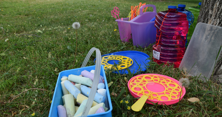  A bucket of sidewalk chalk and an assortment of bubbles sit in the grass near a tree.