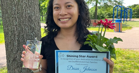  an Asian-American woman, Dessa, is standing at a park. She is holding a Shining Star certificate, glass award, and gerbera daisies.