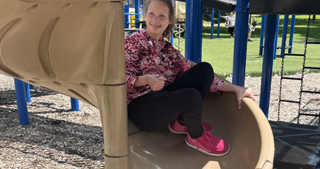 A teenage girl with light skin is coming down a tan spiral slide. She is wearing bright pink shoes and a colorful shirt and smiling.