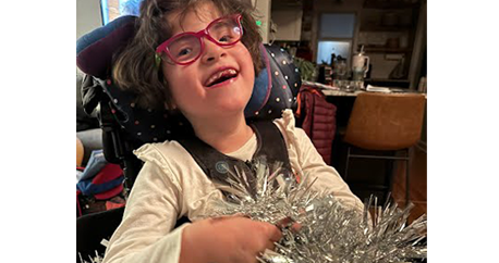 An adolescent girl with light skin and pink glasses sits in her wheelchair holding a silver pom pom and smiles.